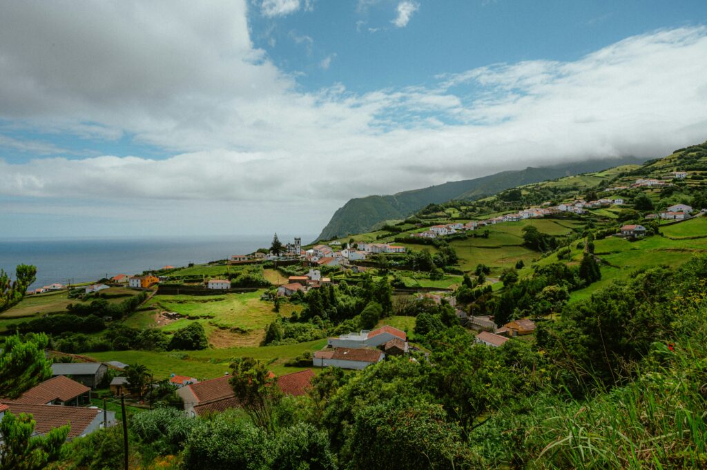 A panoramic landscape view of a lush green hillside village on São Miguel Island in the Azores, Portugal, with clustered white houses featuring red-tiled roofs nestled among terraced fields and vegetation, a prominent church tower, dramatic misty cliffs, and the Atlantic Ocean in the background under a partly cloudy sky.