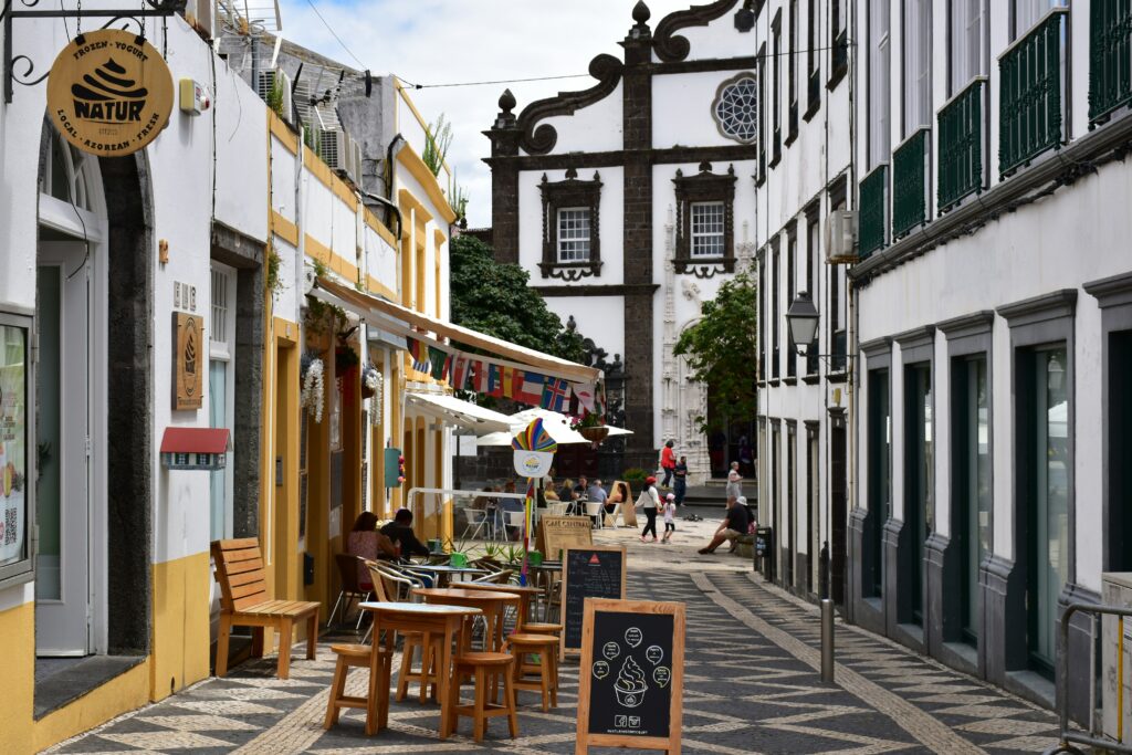 A vibrant narrow cobblestone street in a historic Azores town lined with white and yellow colonial-style buildings featuring ornate dark basalt trim and a tall church facade with a rose window, featuring an outdoor café with wooden tables, chairs, international flags, umbrellas, potted plants, pedestrians, and a "Natur" shop sign under a partly cloudy sky. Azores Travel Guide