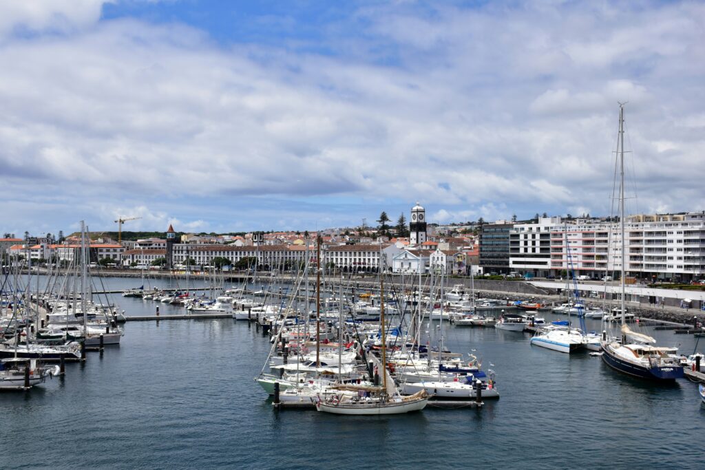 A panoramic view of a bustling Ponta Delgada marina in the Azores filled with numerous moored sailboats and yachts on calm blue waters, set against a historic waterfront in Sao Miguel, Azores, lined with white buildings featuring colorful striped facades, yellow structures, and a distinctive lighthouse on a hill, under a partly cloudy sky. Azores Travel Guide