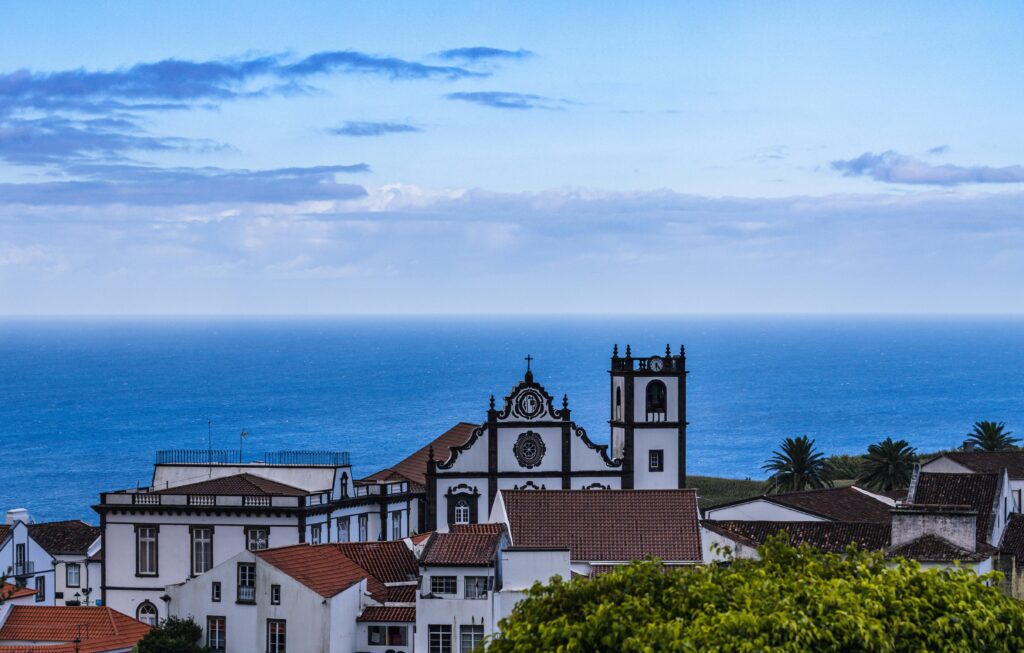 A scenic aerial view of a picturesque Portuguese coastal village featuring a prominent white church with black-trimmed Baroque architecture, a clock tower, and twin bell towers, surrounded by red-tiled rooftops, lush green vegetation, and palm trees, overlooking the deep blue Atlantic Ocean under a partly cloudy sky. Azores Travel Guide
